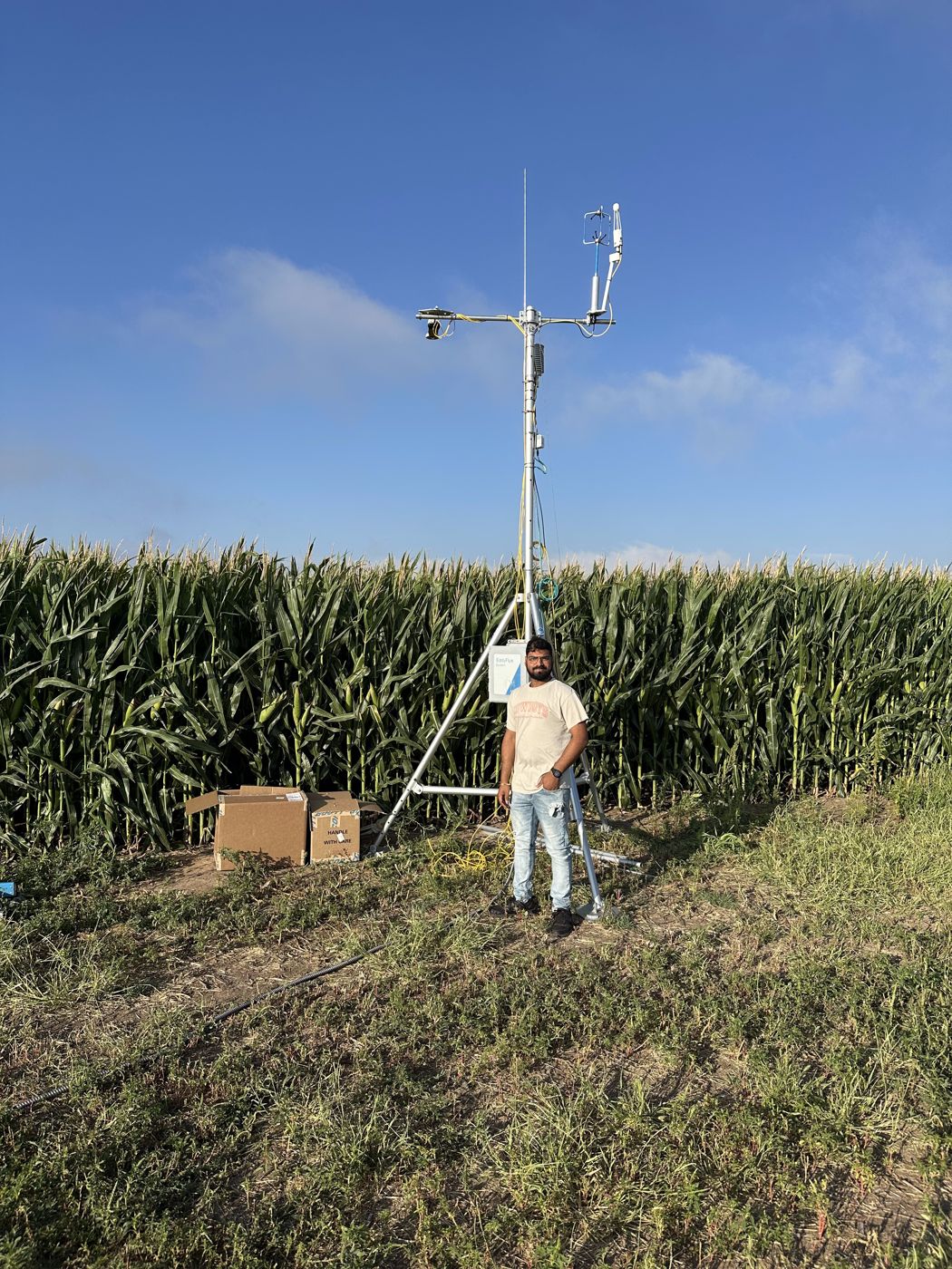 Abinava Yeshwanth KJ standing beside an eddy covariance flux tower at the edge of a corn field